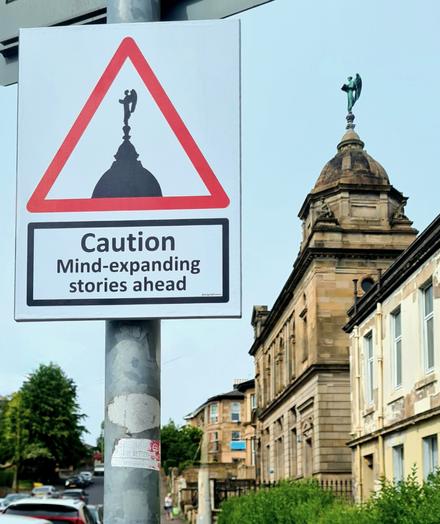 A sign on the road up to a Glasgow public library which reads Caution Mind-expanding Stories Ahead.
