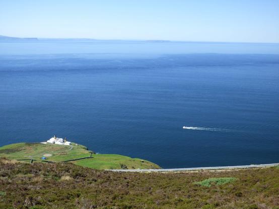 An area of rough heather covered grounds slopes down to a grassy promontory with a narrow winding road, stone walls, and a circular helipad. On the blunt point stands a cluster of white flat roofed buildings with a short yellow lighthouse in the middle. In the middle distance a small vessel passes from right to left, discernable mostly but its white wake on the deep blue sea. The sea is calm under a pallet blue sky and the scene is bathed in bright hot sunshine.