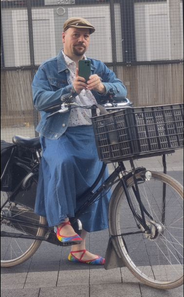 Selbstporträt in spiegelnder Scheibe. Ich sitze auf meinem Fahrrad mit einem Fuß auf dem Boden. Ich trage eine blaue Jeansjacke, einen langen weiten blauen Jeansrock, ein weißes Hemd mit bunten Punkten, bunte Ballerinas in Regenbogenfarben und roten Kreuzgummis, und eine mittelbraune Schirmmütze. Das Hemd ist in den Rockbund gesteckt. Das Fahrrad ist ein schwarzes Hollandrad mit Schwanenhals-Rahmen, Kettenschutz und Speichenschutz.