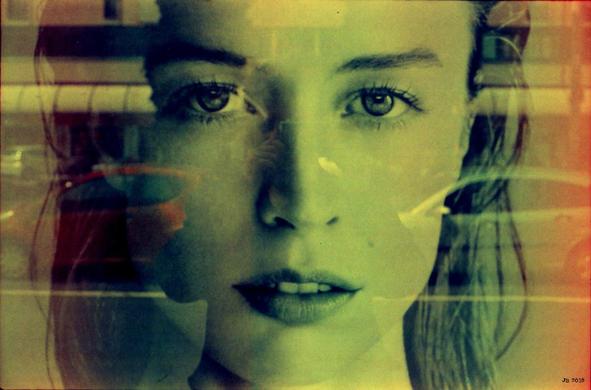 Portrait einer jungen Frau mit Spiegelungen, die ihr Gesicht teilweise überlagern, darunter eine Ladenzeile und davor geparkte Autos. Verfremdete Farben im Spektrum grün-rosa durch den verwendeten Film. Olympus 35SP auf Lomochrome Purple.