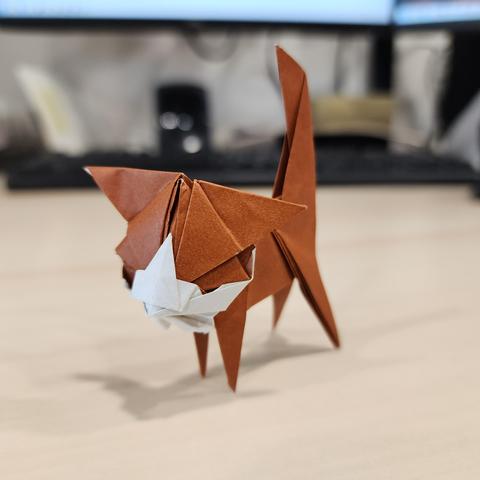 Un chaton en origami, plié dans un papier brun et blanc. Il est posé sur un bureau, on distingue un clavier en arrière-plan. Le chaton a des jambes très simples, mais une tête bien détaillée.