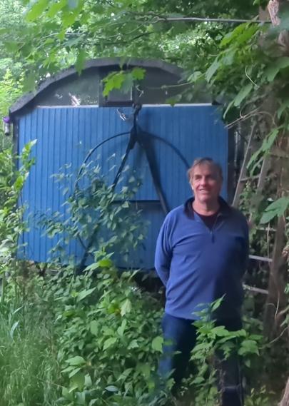 Mann im blauen Pulli und in blauer Jeans vor der Rückseite eines blauen Bauwagens. Hinter ihm ein großes A im Kreis (Anarchisten A). Drumherum grüne Pflanzen.