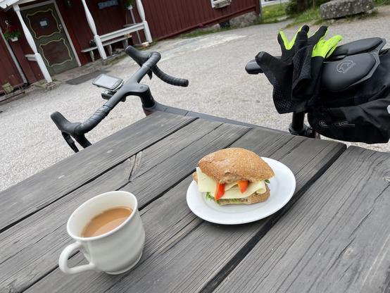 Coffee, sandwich and a bike leaning on the table on the other side of the table.