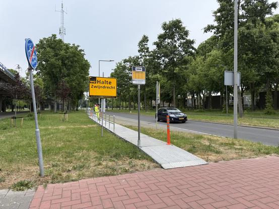 Temporary bus stop in a grass field. An aluminium platform is visible. A yellow sign says “Halte Zwijndrecht” and “Doe voor vertrek de Reisplanner check!”. Road visible right.