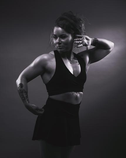 The image is a black-and-white photograph of a person striking a pose. The figure is muscular, wearing a sleeveless top and a skirt. The person has tattoos on their arms and is posing with one arm bent over their head and the other on their hip. The lighting highlights their physique against a subtle background.