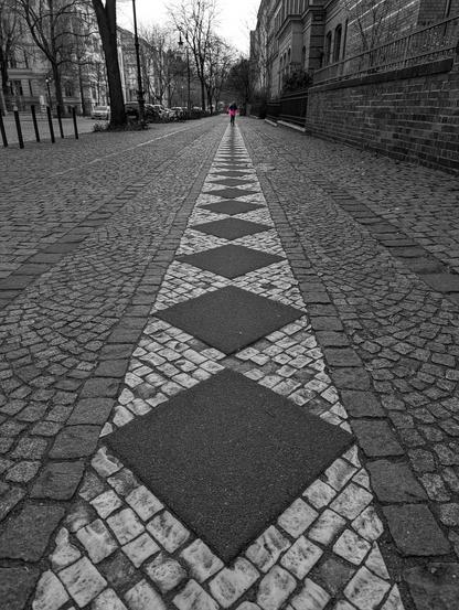 The image is a black and white photograph of a cobblestone pavement next to a road (visible on the left). The pavement stretches into the distance, creating a strong sense of perspective. A child wearing a bright pink winter jacket is running towards the viewer. The cobblestones on the pavement are arranged in a pattern, with a central strip of diamond shapes running along its centre. Buildings line the right side of the pavement, and bare trees are visible on the left, next to the road.