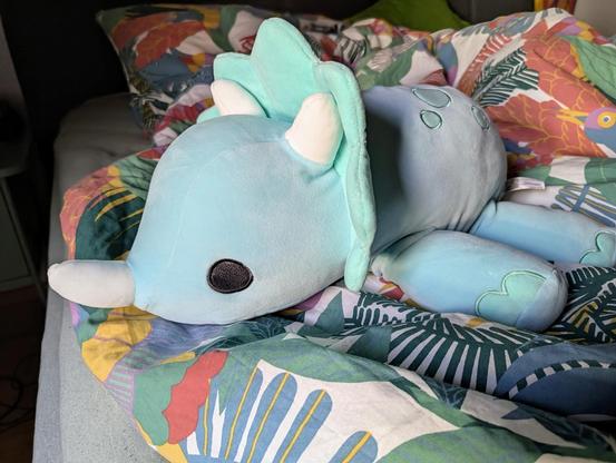 Ein pastellblaues Triceratops-Kuscheltier auf einer bunten Bettdecke.