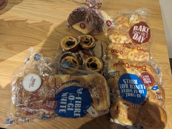 A bunch of bakery items in Bakers Delight bags. It features a loaf of bread, 6 X bread rolls, 6 X mini cheese and bacon rolls, 4 x Portuguese tarts and 4 x scones.