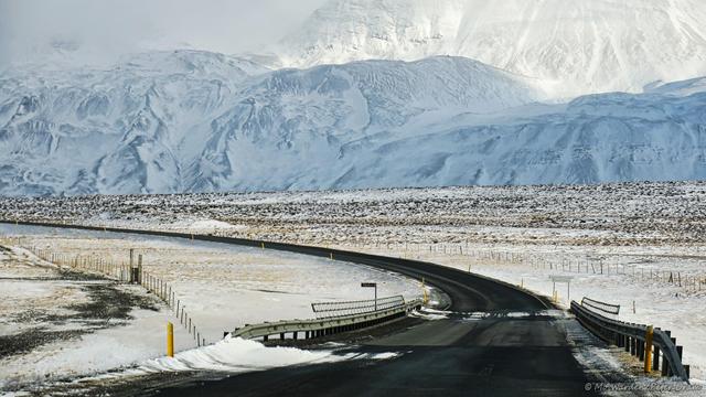 A colour photo of a landscape. The viewpoint is from the front seat of a car travelling along a black tarmac road. Just ahead is a bridge with traffic barriers on either side and a signpost saying "Gljúfurá". The road is curving towards the foot of a mountain covered in snow and ice. The land on either side of the road is also covered.
