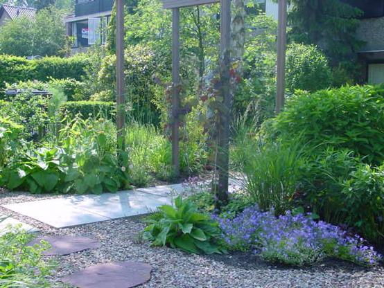 Another view of the front garden. Now from the street. In the foreground a gravel path with flagstones. Behind that from left to right a straight path of large gray concrete tiles. In the middle is a pergola with 4 poles that stand on either side of the tiled path. Around the paths green shrubs and plants. In the background even more green shrubs in the gardens of the neighbors.