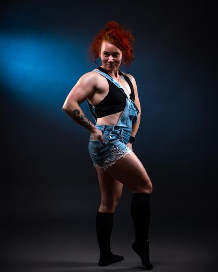 The image shows a person posing confidently. They have red hair and are wearing denim overalls, a black top, and black knee-high socks. The background has a dark setting with a blue light creating a spotlight effect.