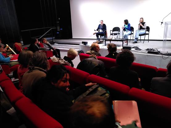 Amphithéâtre avec des sièges rouges. Estrade avec 3 personnes à la tribune