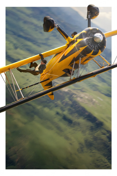 Foto enmarcada en filo blanco. Se ve una avioneta amarilla volando boca abajo y Tom Cruise enganchado en una de sus alas