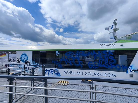 Zu sehen ist die Steuerbordseite der Warnowfähre zwischen Gehlsdorf und Kabutzennhof. Über Nacht haben Vandalen mit blauer Farbe fussballbezogene "Grüße" auf Scheibe und Aussenwand gesprüht. Im hinteren Teil des Schiffes sind unleserliche Tags geschmiert.
