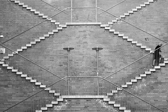 Schwarz-Weiß-Foto. An einer Backsteinwand sind mehrere Treppen und Geländer angebracht. sie ergeben in der Frontansicht ein symmetrisches geometrisches Muster und wirken gar nicht wie dreidimensionale Treppen. In der Bildmitte sind zwei ebenfalls symmetrische Laternen. Am rechten Bildrand steift ein Mann die Treppen hinauf und unterbricht damit die Symmetrie.