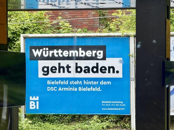 Das Bild zeigt ein großes blaues Werbeplakat mit dem Text "Württemberg geht baden. Bielefeld steht hinter dem DSC Arminia Bielefeld." Der Hintergrund des Plakats ist blau, und der Text ist in weißer und schwarzer Schrift dargestellt. Links unten ist das Logo von Bielefeld Marketing mit den Buchstaben "BI" und einem weißen Wappenturm zu sehen. Darunter steht "Bielefeld Marketing Für dich. Für die Stadt. www.bielefeldJETZT." Im Hintergrund sind grüne Pflanzen und ein rotes Backsteinhaus zu erkennen. Das Plakat ist an einem Metallgestell befestigt und befindet sich in einer offenen Umgebung, möglicherweise an einer Haltestelle oder einem öffentlichen Ort.