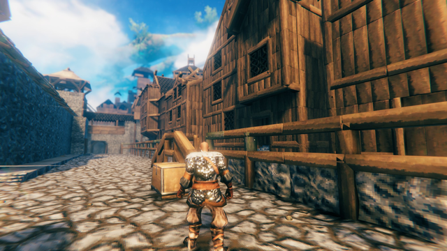 Valheim Screenshot: Eine Straße, auf deren rechter Seite einige Holzhäuser zu sehen sind und auf der linken Seite eine Stadtmauer oder ähnliches. Die Straße läuft auf ein Tor zu. Im Hintergrund sind höhere Türme erkennbar.