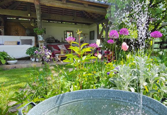 Das Bild zeigt einen idyllischen Garten mit einer Vielzahl von Blumen und Pflanzen im Vordergrund. Vorderste Position hat ein großes, runder Wasserbehälter, aus dem Wasser spritzt und in den Behälter fällt. Die Blumen sind in verschiedenen Farben und Größen angeordnet, darunter auffällige violette Alliumsblüten und rosa Tulpen. Im Hintergrund ist ein gemütlicher Gartenbereich mit einem Holzgestell und einem  Dach zu sehen. Darunter ein Sofa mit Kissen und ein Tisch, sowie weitere Pflanzen in Topfpflanzen. Die Umgebung wirkt gepflegt und einladend, mit einer harmonischen Mischung aus Natur und gemütlicher Einrichtung.