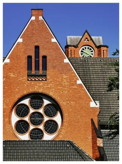 Das Bild zeigt in Teilen die Architektur einer Kirche. Der Vordergrund wird von einer Giebelfront aus rotem Backstein dominiert. Die Giebelfront hat ein großes, kreisförmiges Fenster, das in mehrere kleinere runde Fenster unterteilt ist (Rosenfenster). Darüber befinden sich drei schmale, vertikale Fenster. Hinter dieser Giebelfront ist ein höherer Turm sichtbar, der ebenfalls aus Backstein besteht und zur Kirche gehört. Unter dem Giebel befindet sich eine große Uhr.

**Hintergrundumgebung:**

*   Der Hintergrund ist ein klarer, blauer Himmel. Es gibt keine Wolken oder andere Elemente, die vom Gebäude ablenken würden.

**Farbdetails:**

*   Die Hauptfarbe ist ein warmer, roter Backstein. Die Farbe variiert leicht aufgrund von Licht und Schatten, was der Fassade Tiefe verleiht.

*   Die Fenster sind dunkel, vermutlich weil sie nicht beleuchtet sind oder stark reflektieren.

*   Das Dach des Gebäudes und des Turms ist dunkelgrau oder schwarz.

*   Der Himmel ist ein kräftiges Blau.

**Emotionaler Ausdruck:**

*   Das Bild vermittelt ein Gefühl von Stabilität und Dauerhaftigkeit, vermutlich aufgrund der soliden Backsteinarchitektur.

*   Es hat auch einen Hauch von Erhabenheit und spiritueller Bedeutung, da Kirchen oft eine solche Wirkung haben.

*   Die klaren Linien und die symmetrische Anordnung der Elemente tragen zu einem Gefühl von Ordnung und Harmonie bei.

**Detaillierte Nahaufnahmen:**

*   Die Textur des Backsteins ist sichtbar, was dem Bild eine taktile Qualität verlei