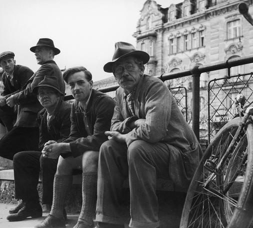 Vor einem Geländer sitzen drei Männer, die in die Kamera blicken, dahinter lehnen zwei andere. Rechts das Rad eines Fahrrads. Im Hintergrund eine Häuserfassade. Wien, 1938. Nachweis von wiki commons folgt.