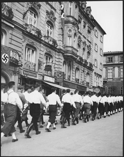 Österreich, Salzburg: Menschen; Männer mit Hakenkreuz-Armbinde maschieren durch die Strassen. Fotografie zeigt Männer, die abgewandt von der Kamera nach rechts marschieren. Hinter ihnen eine Hauswand mit Geschäften und einer Hakenkreuz-Fahne. Nachweis von wiki commons folgt.