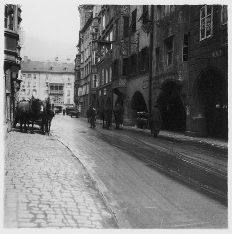 Die Fotografie zeigt eine eher leere Straße in Österreich, Innsbruck: Häuserzeile; Strasse in Innsbruck mit Pferdefuhrwerk. Nachweis von wiki commons folgt.
