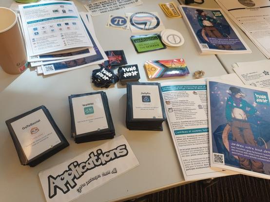Une photographie de la table du stand YunoHost, avec au premier plan des cartes YugiHost promouvant quelques applications du catalogue YunoHost, ainsi qu'en libre accès des flyers et stickers