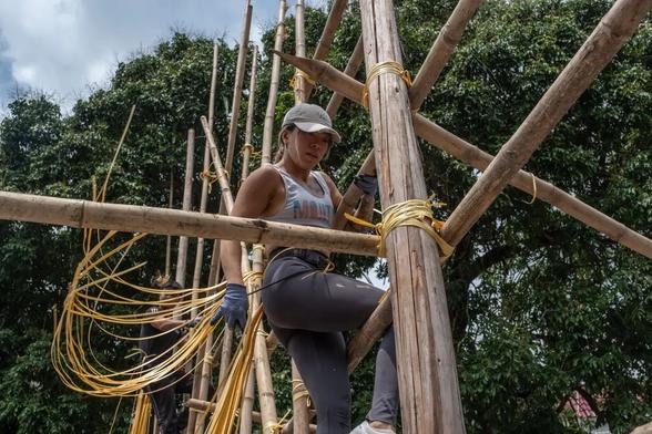 A muscular Chinese woman wearing a cap standing on a bamboo scaffolding as she ties it together, with trees in the background.

Photograph by Lam Yik Fei, from:

https://www.nytimes.com/2025/05/24/world/asia/hongkong-bamboo-scaffolding.html