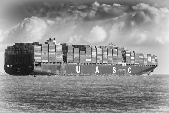 This Is A High Contrast Black And White Seascape Photo In Landscape Format. Filling The Lower Third Of The Frame Is A Body Of Calm Sea Water. Just Below The Horizon Is An Oblique View And Seen From The Stern, Of A Very Large Container Ship Underway From Left To Right. The Ship Stretches From The Left To Right Margins. Above Deck Are Thousands Of Stacked (Nine High In Places) Forty Foot Shipping Containers. The Containers Are Of Many Colours Rendered Into Different Tones In Black And White. The Low Sun Is Illuminating The Starboard Side Of The Weathered Hull And Containers Though The Stern Is In Shadow. The Hull Is Hull Is Black With 'UASC' In Vast White Bold Letters, The Initials Of The 'United Arab Shipping Company'. Just Visible On The Stern Are The Vessel's Name 'SAJIR' And 'Majuro' The Port Of Registry. The Hull Is Black And The Superstructure, White, As Is Common On Merchant Shipping. Above The Ship And In the Near Distance Is A Cloudy And Stormy Sky Made Up Of Cumulus And Developing Cumulonimbus Clouds. 
The 368 Metre (1200ft) Sajir Is Leaving The Port Of Southampton And Threading Her Way Through The Deep Water Channel Towards The English Channel (2019).