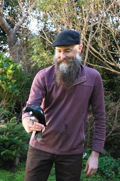 A photo of Chris, a Level 3 bird bander, with one of the 15 new tūī caught today in a mist-net and banded before being released. Chris is holding the tūī carefully in one hand and he's smiling.