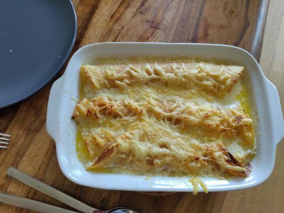 Auf dem Bild sieht man ein Gericht, das in einer weißen Auflaufform serviert wird. Es handelt sich um gefüllte Teigrollen, die mit Käse überbacken sind. Der Käse ist geschmolzen und hat eine goldbraune Farbe. Neben der Auflaufform liegt ein grauer Teller sowie Besteck (Gabel und Löffel) auf einem Holztisch.
