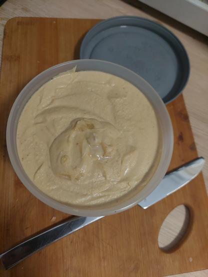 Ein rundes Plastiktöpfchen mit frischem Hummus steht auf einem Holzbrettchen. Daneben liegen Tafelmesser und Deckel (nicht ganz im Bild)
