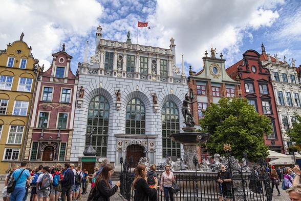 We're in a wide and busy pedestrianised area of a city with houses and other buildings along that look to be centuries old. Buildings in various colours with four floors and stepped and sometimes curved gables are either side of a wider, more impressive building in pale blue, large, stone bricks, with three enormous arched window fronts rising up three floors and an ornate doorway in the centre. In the pedestrian area there is a fountain with a figure with trident atop it and many people are walking past or admiring the sights.