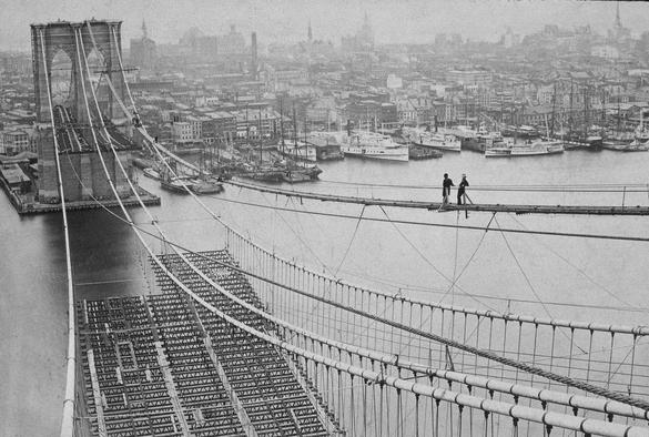 The Brooklyn bridge under construction. The towers have been built and the suspension cables hung. The roadway is still just girders hanging in air. Two workmen are lounging on the cable.