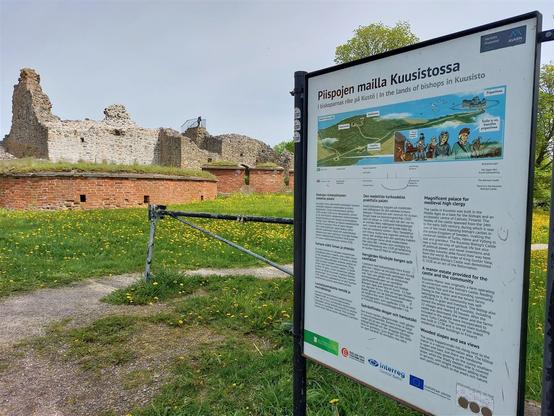 Yksi opaskyltti piipojen maille Kuusistossa, taustalla rauniot