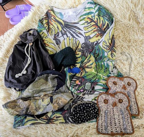 Auf einer flauschigen weisen Decke liegen Flohmarktsachen. Ein Pullover mit Blättermuster, ein schwarzer Stoffrucksack, ein gestricktes grünes Stirnband, ein Seidentuch, 2 gestrickte Eulentopflappen, ein kleiner schwarzer Geldbeutel und Schmuck.