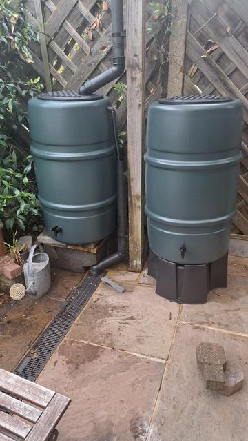Two large green water butts. One on a concrete plinth and one on a plastic stand. The one on the left has the downpipe feeding into it and has an overflow leading into the patio drain