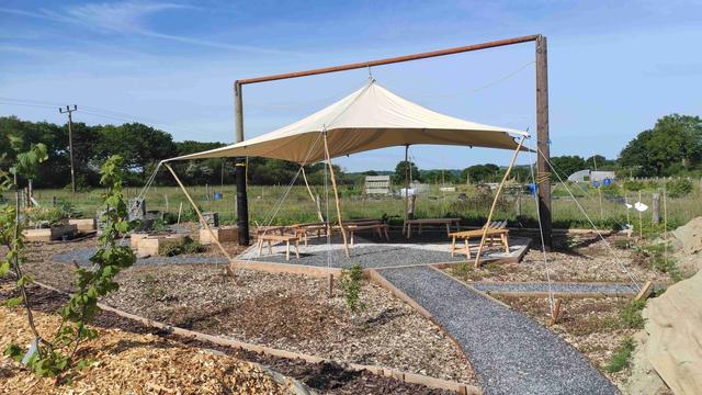 Canvas shelter supported from crossbar between 2 telegraph poles in a field by an allotment and in midst of half planted garden
