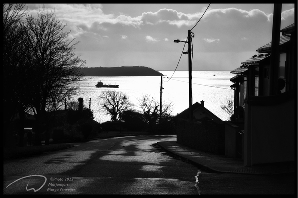 View over an abandoned street sloping down with houses on the right and trees on the left, a power pylon with cables and views over the atlantic ocean with a freighter in the distance.