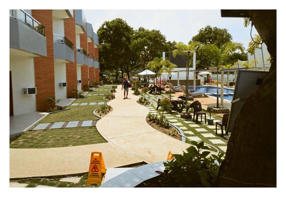 An outdoor area of Crown Of The Orient Beach Resort in Gloria, Oriental Mindoro, Philippines.

On the left, there's a multi-story building with a modern design, featuring balconies and a combination of white and reddish-brown (brick-like) facades. In front of the building, there's a well-maintained lawn with stepping stones and landscaped garden beds.

A curved pathway made of light-colored material winds through the scene, leading towards a swimming pool area visible further back on the right. Along this pathway, there is a "caution: wet floor" sign at the bottom left of the picture.

The area around the pool features lounge chairs, umbrellas, and what looks like a poolside bar or seating area. There are numerous palm trees and other lush green trees.

In the midground, a person is walking along the curved pathway away from the viewer. - Google Gemini