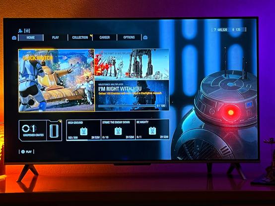 A television screen displaying a gaming interface for a multiplayer game, featuring options like "Quick Match" and "I'm Right With You." The screen includes various game modes and milestone tracking, with a robotic character in the corner. The background is softly lit.