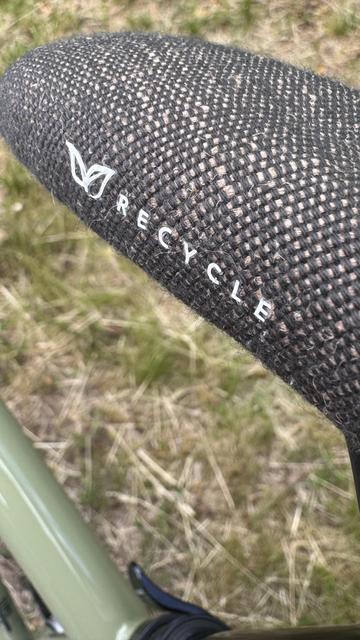 Eine Nahaufnahme eines Fahrradsitzes mit einem strukturierten schwarzen Bezug, mit dem Wort "RECYCLE" und einem Blatt-Emblem vor einem grasigen Hintergrund.