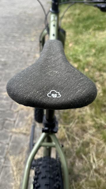 Nahaufnahme eines strukturierten schwarzen Fahrradsattels mit einem weißen Wolkenlogo. Das Fahrrad befindet sich auf einem Weg mit sichtbarem Gras im Hintergrund.