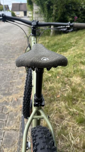 Eine Nahaufnahme eines Fahrradsattels und Rahmens, mit einem verschwommenen Fokus auf den Lenker. Das Fahrrad verfügt über grobstollige Reifen, und die Einstellung umfasst einen gepflasterten Weg und Gras im Hintergrund.