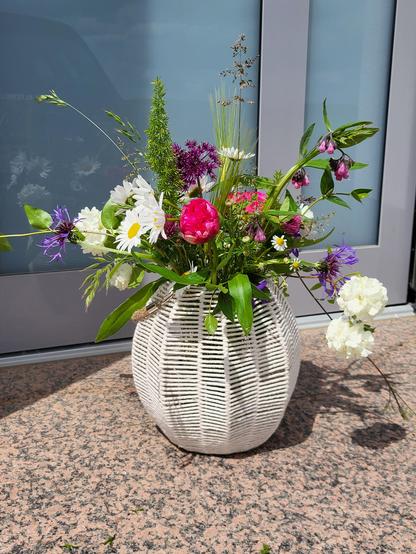 Das Bild zeigt eine farbenfrohe Blumenarrangement in einem weißen, geflochtenen Vasen, die auf einem Granitboden vor einer Glasfront steht. Die Blumen bestehen aus einer Vielfalt von Pflanzen, darunter weiße und rote und lila Blumen, sowie verschiedene Gräser und Blätter. Die Blumen sind in einer natürlichen, wilden Art arrangiert, was ihnen ein lebendiges und frisches Aussehen verleiht. Der Hintergrund besteht aus einem grauen Fenster, das das Licht reflektiert und einen modernen Kontrast zur natürlichen Blumenarrangement bildet. Die Vasenstruktur ist geometrisch und verleiht dem Ganzen eine elegante Note.