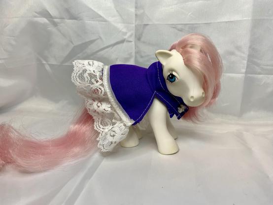 A white pony figurine wearing a purple outfit with lace trim. The pony has long pink hair and blue eyes. The background is plain white.