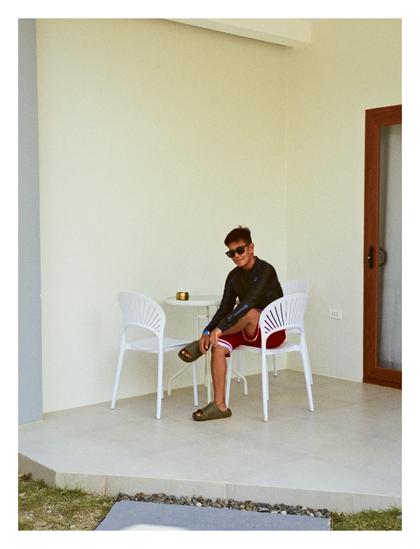 My aunt, with her glasses on, sitting on a white chair at a small, round table on a porch in a resort's accommodation building.