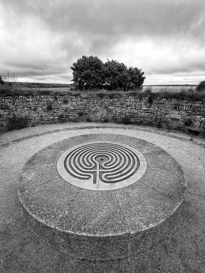 A large circular stone. The flat top has a metal plate with the map of a maze on it. Around the stone is a curved stone wall. Trees and a lake can be seen behind the wall in the distance.