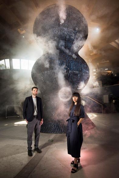 image/jpeg a man and a woman stand in a large space in front of a giant blue sculpture shaped like three stacked balls, which is emitting large vapour rings. Photo from Studio Swine.
