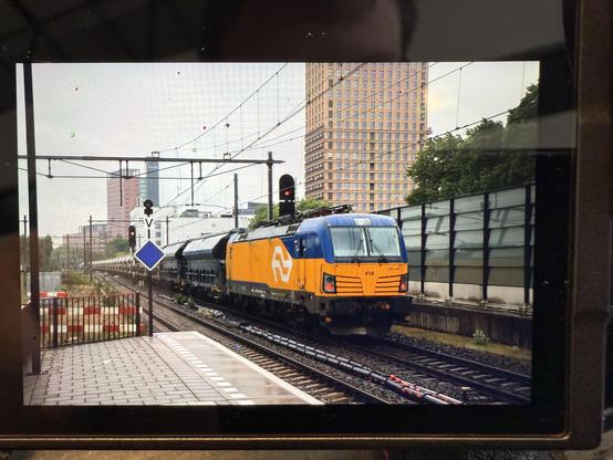 A photo of a camera screen displaying a photo. Visible is a yellow-blue locomotive with red (end) signals. Behind it a cargo train. A platform is visible in front and some high rise buildings in the distance.